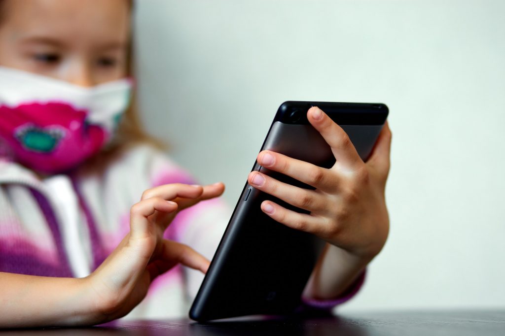Young child girl at home, wearing a protection mask and gaming on a tablet, Coronavirus quarantine.